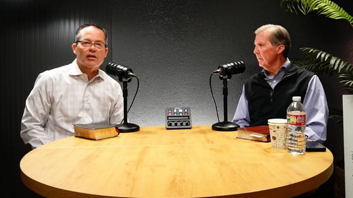 Man seated to the left behind a light wooden table in a button up and a podcast mic. Man in a vest seated to the right behind a podcast mic.