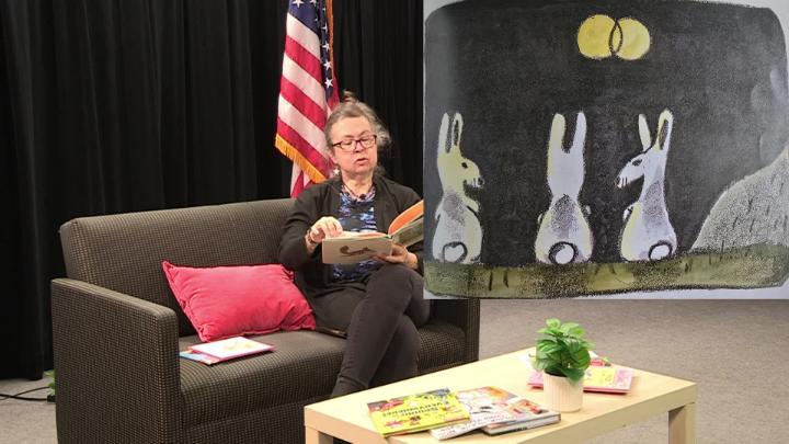 Woman seated on a couch reading a book. American flag in the background. On right, a picture of the book page with three white rabbits