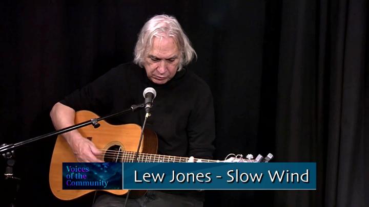 Black curtain background. Older man dressed in black seated behind a mic playing a guitar. Words at the bottom: "Lew Jones - Slow Wind"
