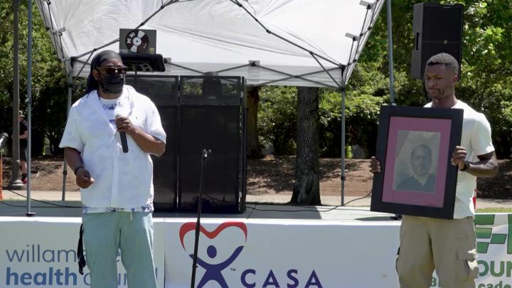 African American man in a white shirt standing with a mic to the left. African American man holding a picture standing to the right.