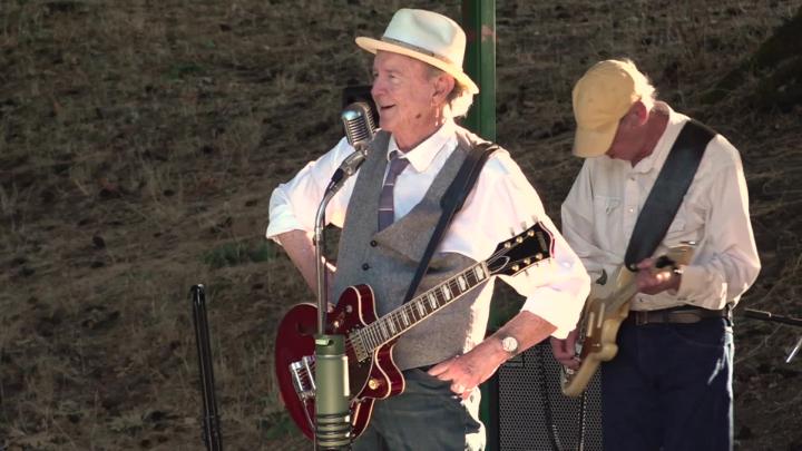 Outside - Man in a button up vest in front of a mic, holding a guitar