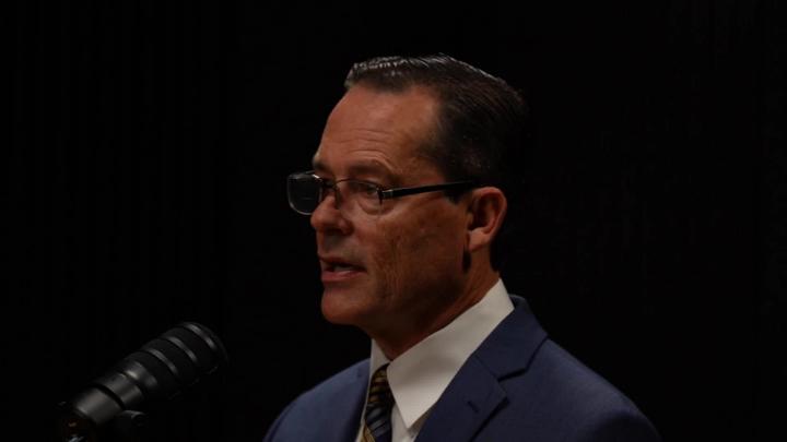 Man in a suit and glasses facing the left in front of a podcast mic