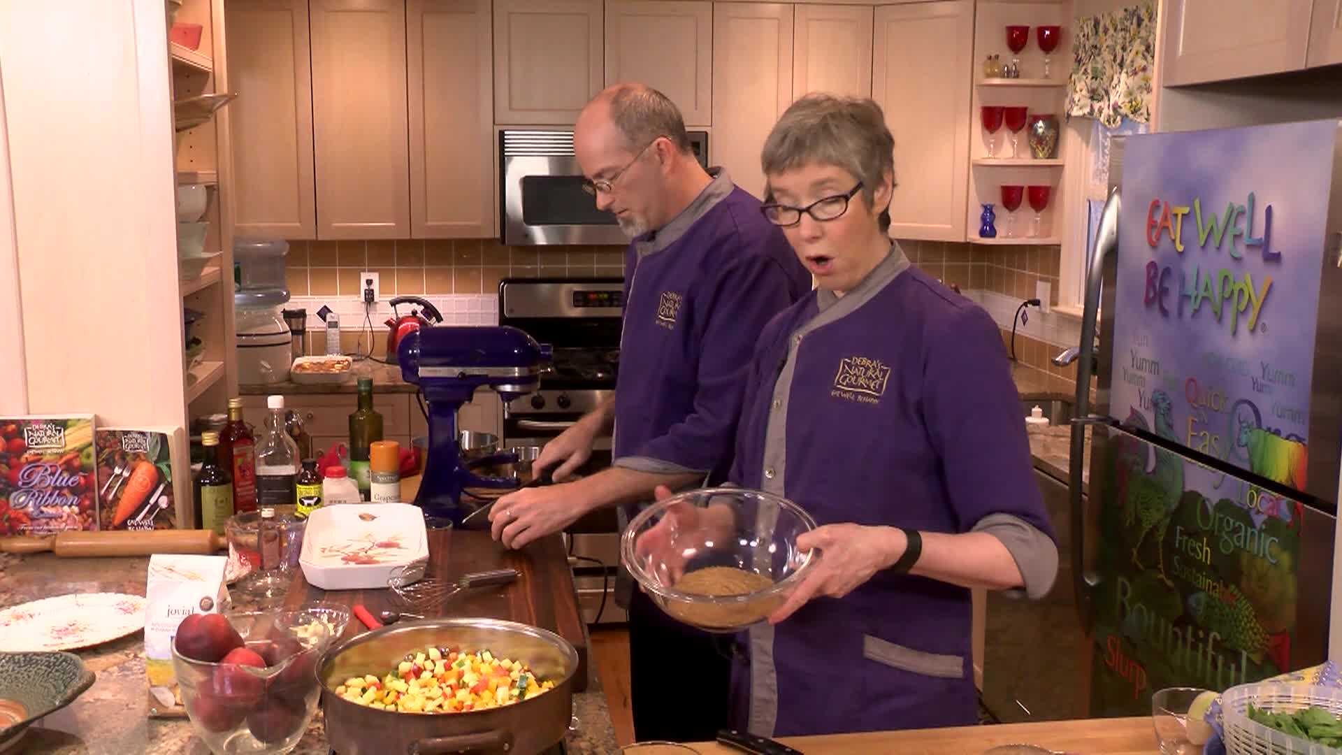 Kitchen setting. Woman with glasses and purple chef jacket to the middle right. Man with glasses and purple chef jacket to middle left preparing food.