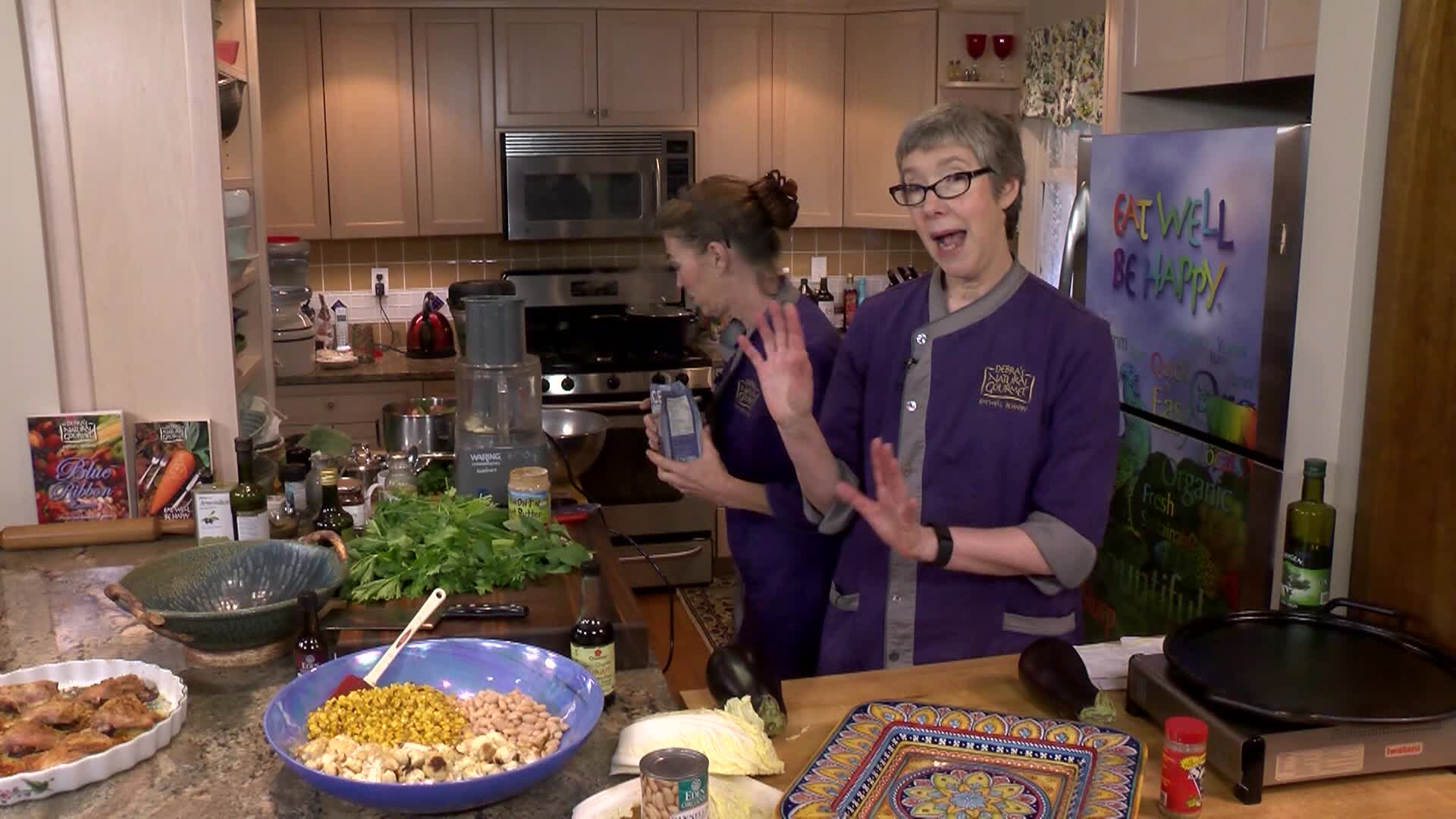 Woman in purple on the left in a kitchen cooking. Woman in purple in the center prepping food.